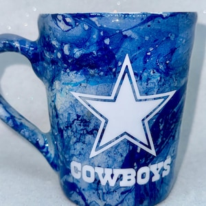 May include: A blue and white ceramic mug with a white star and the word "COWBOYS" in white lettering. The mug has a marbled blue and white design.