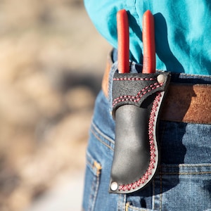 Może przedstawiać: Czarny skórzany futerał na narzędzia z czerwonymi szwami, trzymający dwa narzędzia z czerwonymi uchwytami. Futerał jest przymocowany do brązowego skórzanego paska noszonego z niebieskimi dżinsami. Tło jest rozmyte.