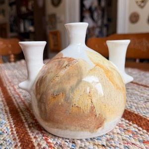 May include: A white ceramic vase with three spouts. The vase has a rounded body with a textured, earthy design in shades of brown, tan, and yellow. The spouts are cylindrical and extend upwards and outwards from the body.
