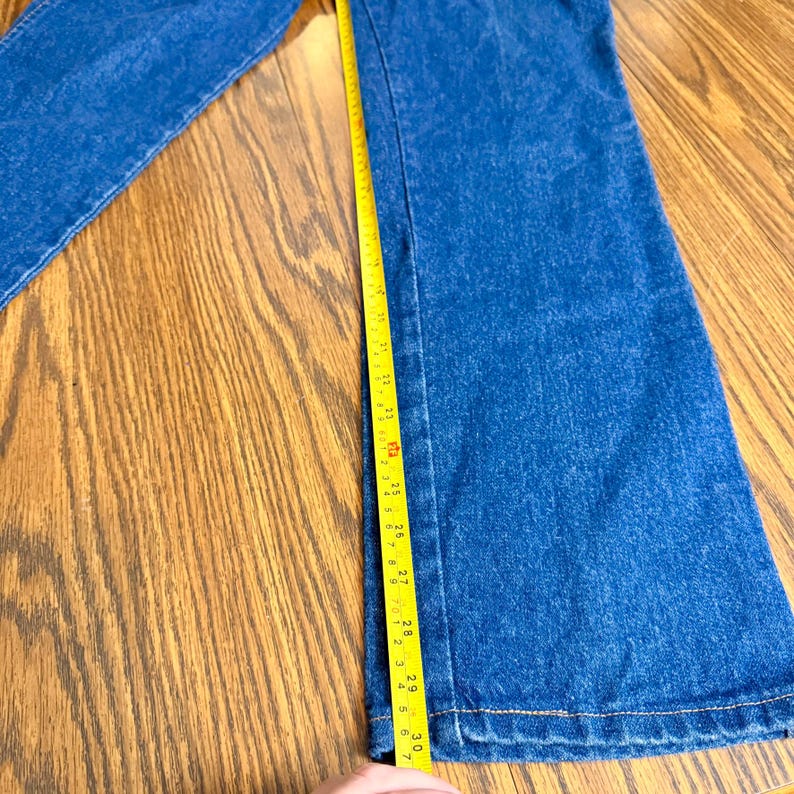 May include: Blue denim jeans with a flared leg, measured with a yellow tape measure. The jeans measure approximately 30 inches in length from the waist to the hem.