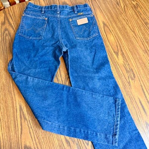 May include: Blue denim jeans with a classic five-pocket design. The jeans feature the Wrangler logo on the back pocket and a leather patch. The jeans are made of a durable denim fabric and have a straight leg cut.