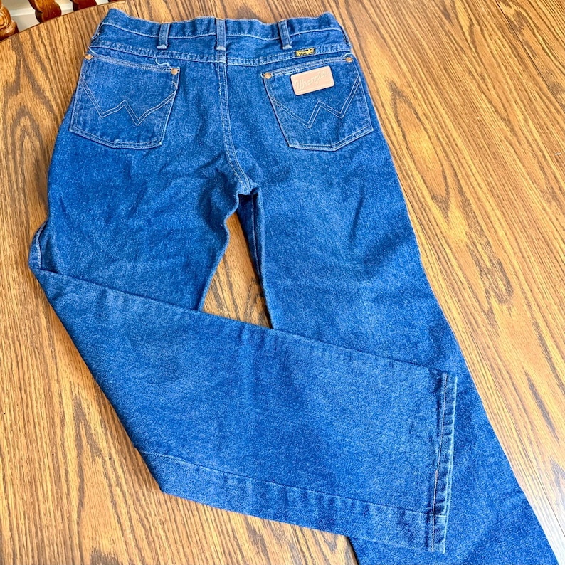 May include: Blue denim jeans with a classic five-pocket design. The jeans feature the Wrangler logo on the back pocket and a leather patch. The jeans are made of a durable denim fabric and have a straight leg cut.