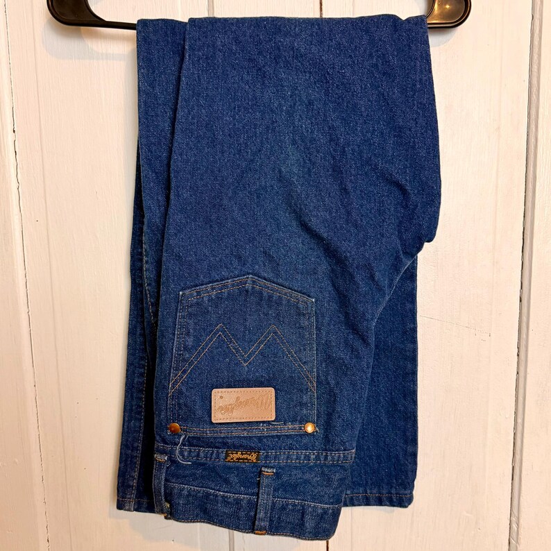 May include: Dark blue denim jeans hanging on a black hanger. The jeans have a classic five-pocket design with a brown leather patch on the back pocket. The Wrangler logo is visible on the patch and the front pocket.