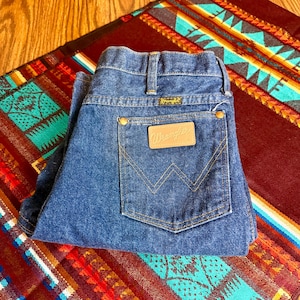 May include: Blue denim Wrangler jeans folded on a colorful patterned textile. The jeans feature the Wrangler logo on the back pocket and waistband. The jeans have a classic five-pocket design with brass-colored rivets.