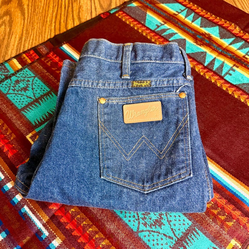 May include: Blue denim Wrangler jeans folded on a colorful patterned textile. The jeans feature the Wrangler logo on the back pocket and waistband. The jeans have a classic five-pocket design with brass-colored rivets.