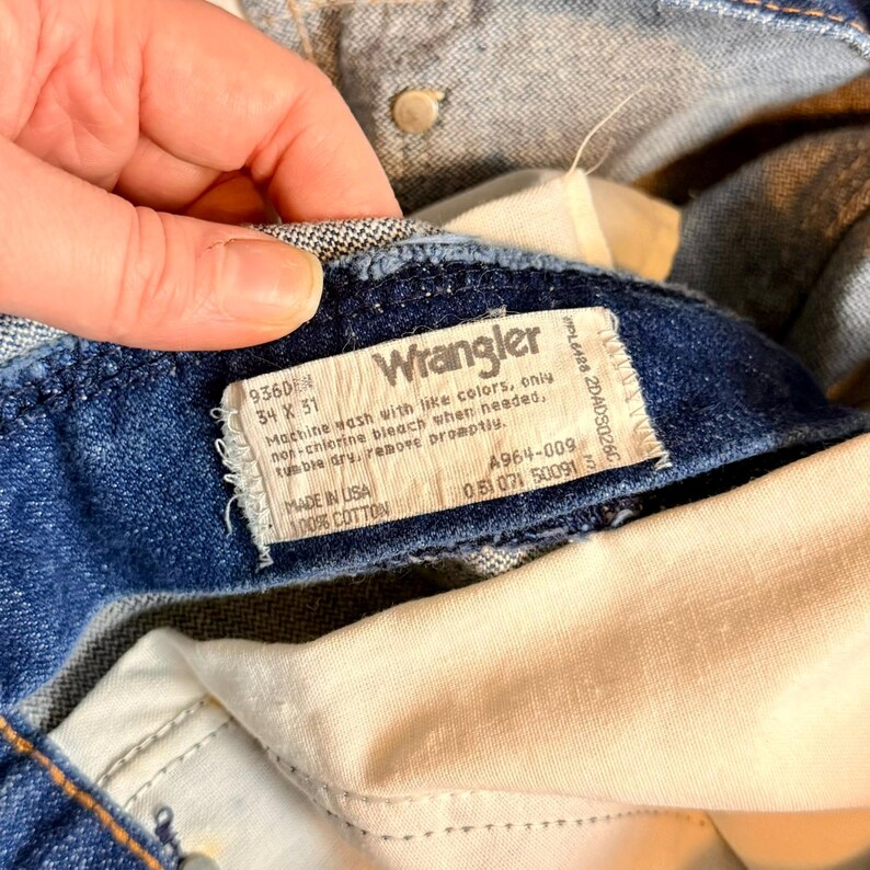 May include: Close-up of a pair of blue denim Wrangler jeans. The label reads "Wrangler" and includes washing instructions and size 34 x 51. Made in USA, 100% cotton. The jeans are folded, showing the inside lining.