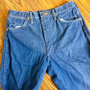 May include: Close-up of blue denim jeans. The jeans have a classic five-pocket design with a button and zipper closure. The jeans have copper-colored rivets and a faded appearance.