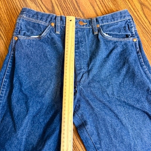 May include: Close-up of blue denim jeans with a ruler placed vertically. The jeans have classic five-pocket styling, a button closure, and copper-colored rivets. The ruler shows measurements in inches.