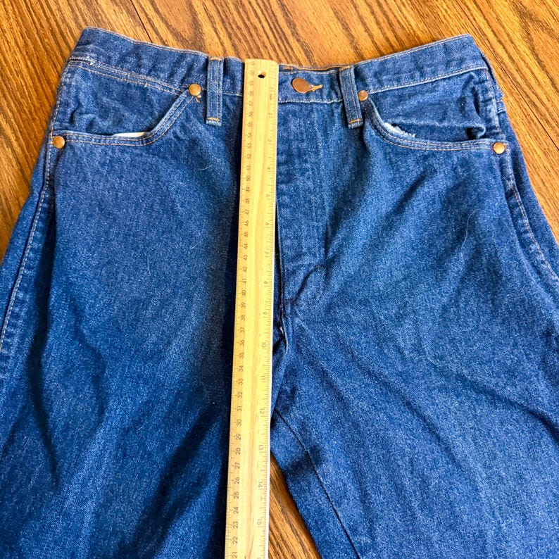 May include: Close-up of blue denim jeans with a ruler placed vertically. The jeans have classic five-pocket styling, a button closure, and copper-colored rivets. The ruler shows measurements in inches.