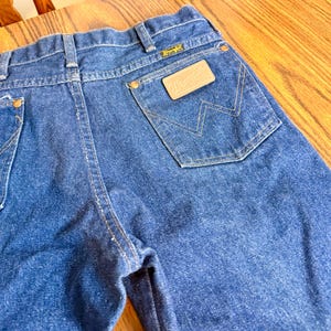 May include: Close-up of blue denim jeans with a Wrangler label on the back pocket. The jeans have a classic five-pocket design with a button and zipper closure. The stitching is a contrasting yellow color.
