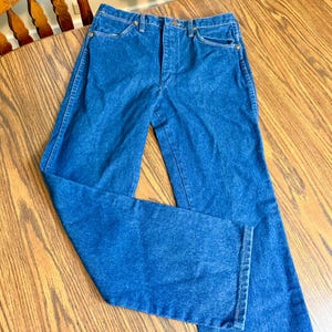 May include: Blue denim jeans with a classic five-pocket design. The jeans have a high waist and a straight leg cut. The denim is a medium wash with visible stitching and brass-colored rivets. The jeans are folded on a wooden surface.