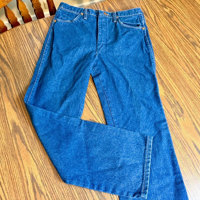 May include: Blue denim jeans with a classic five-pocket design. The jeans have a high waist and a straight leg cut. The denim is a medium wash with visible stitching and brass-colored rivets. The jeans are folded on a wooden surface.