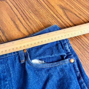 May include: Close-up of a pair of blue denim jeans with a wooden ruler placed across the waist. The ruler shows measurements in inches, with the jeans' waist measuring approximately 30 inches.