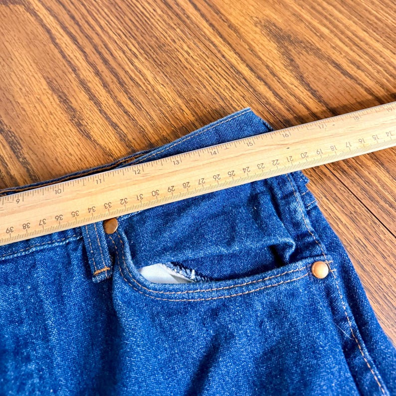 May include: Close-up of a pair of blue denim jeans with a wooden ruler placed across the waist. The ruler shows measurements in inches, with the jeans' waist measuring approximately 30 inches.