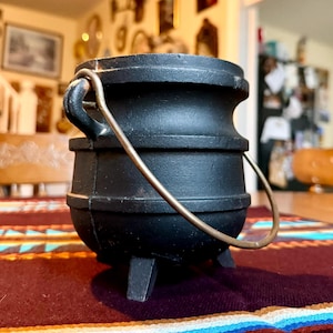 May include: A small, black cast iron cauldron with a metal handle. The cauldron has three small legs and is sitting on a colorful patterned surface. The cauldron is round with horizontal bands around the body.