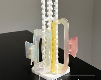 May include: A white vertical hair accessories tower organizer with a scalloped edge design. The stand holds two rectangular hair clips: one iridescent translucent clip and one gradient yellow-to-pink translucent clip. Text reads DADA 3D CRAFT.