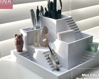 May include: A white 3D-printed desk organizer shaped like a multi-level city house with stairs, featuring compartments for pens and stationery. It includes a small cat figurine on the steps, a brown cat figure, and two crystals. Text reads DADA 3DCRAFT.