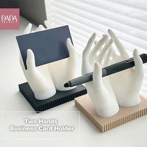 Peut inclure: Deux porte-cartes en forme de mains. L'un tient une carte bleu marine, l'autre un stylo noir. Les supports sont blancs avec des bases noires et beiges. Le texte "Two Hands Business Card Holder" est visible.