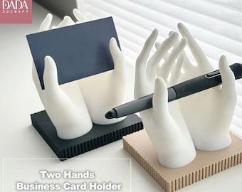 May include: Two hand-shaped business card holders. One holds a navy blue card, the other a black pen. The holders are white with black and tan bases. The text "Two Hands Business Card Holder" is visible.