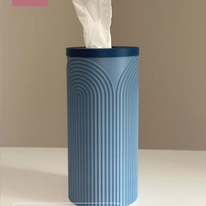 K&ouml;nnte beinhalten: Ein blau-grauer, zylindrischer Taschentuchbox-Bezug mit dunkelblauem Deckel und vertikaler, bogenf&ouml;rmiger Rippenstruktur. Das Produkt ist mit DADA 3DCRAFT und Round Cylinder Tissue Box Cover beschriftet. Ein wei&szlig;es Taschentuch ragt oben heraus.
