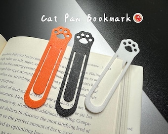 May include: Three cat paw bookmarks in orange, black, and white, each featuring a cutout paw print design at the top. The bookmarks are shaped like elongated paper clips and are resting on an open book. The text Cat Paw Bookmark is visible.