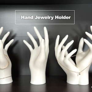 May include: Three white ceramic hands designed to hold jewelry. The hands are posed in different positions, with one hand holding a bracelet, another holding a ring, and the third holding a necklace.