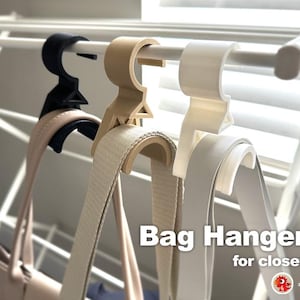 May include: Three plastic bag hangers for a closet. The hangers are beige, white, and black. The hangers are designed to hold the straps of a bag.