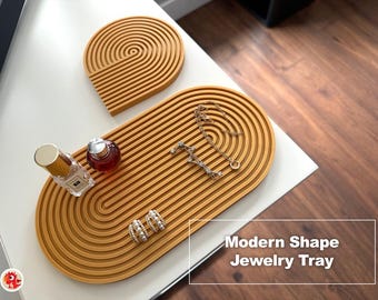 May include: Two mustard yellow trays with a raised concentric ribbed pattern. The larger tray is oval-shaped and holds a small perfume bottle, a gold chain necklace, and a pair of pearl hoop earrings. The smaller tray is a semi-circle shape.
