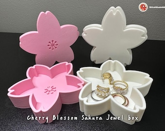 May include: Four pink and white cherry blossom shaped boxes. The boxes are stacked on top of each other. The top box is open and contains several rings. The boxes are labeled "Cherry Blossom Sakura Jewel box".