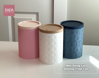 May include: Three cylindrical desktop trash cans with swing lids. The left is pink with an arched pattern, the center is white with a star and circle pattern and a tan lid, and the right is blue with a geometric semi-circle pattern.