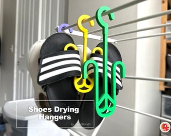 May include: Three plastic shoe drying hangers in green, yellow, and purple, each featuring a hook for suspension and a pronged base to hold footwear. The hangers are displayed on a white rack, with black and white slides hanging from them. Text reads: Shoes Drying Hangers.
