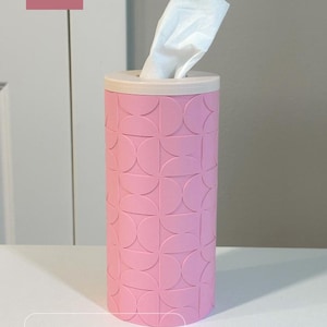 K&ouml;nnte beinhalten: Ein zylindrischer Taschentuchbox-Bezug in Sakura-Pink mit beigem Deckel. Die Au&szlig;enseite zeigt ein sich wiederholendes geometrisches Muster aus Halbkreisen. Ein wei&szlig;es Taschentuch ragt aus der oberen &Ouml;ffnung. Der Text DADA 3D CRAFT und Round Cylinder Tissue Box Cover sind zu sehen.