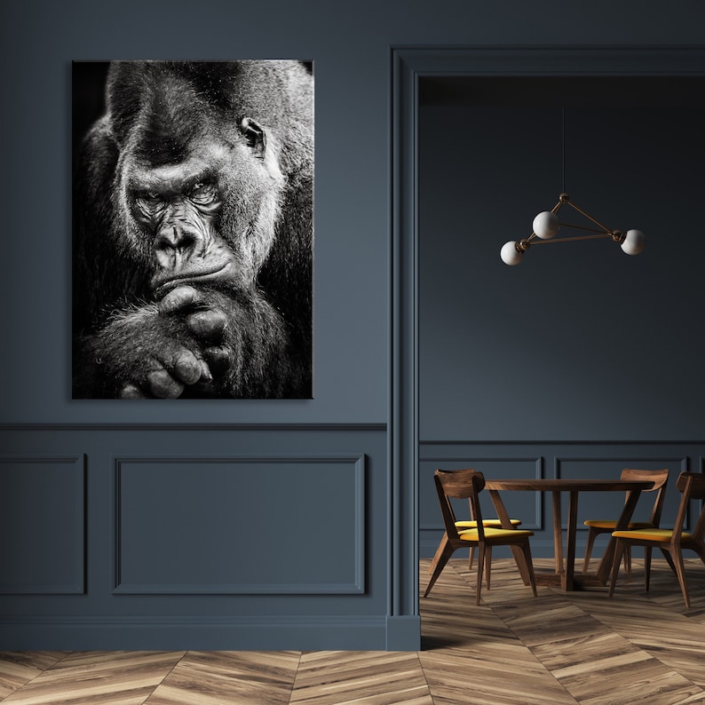 May include: A black and white photograph of a gorilla looking directly at the camera. The gorilla is sitting with its arms crossed and its head resting on its hands.