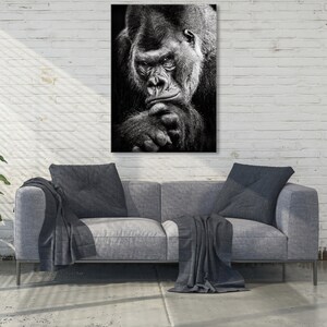 May include: Black and white photograph of a large gorilla looking directly at the camera. The gorilla is sitting with its chin resting on its hand.