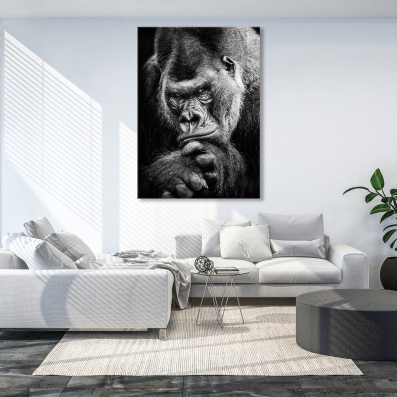 May include: Black and white photograph of a gorilla looking directly at the camera. The gorilla is sitting with its arms crossed and its chin resting on its hand.