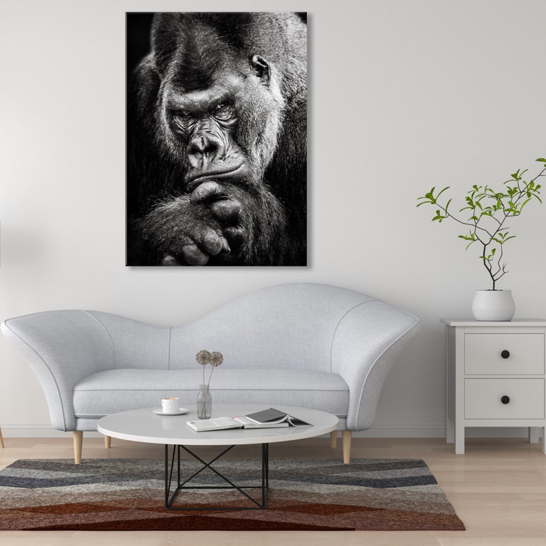 May include: A black and white photograph of a large gorilla looking directly at the camera. The gorilla is sitting with its arms crossed and its head resting on its hand. The gorilla's fur is thick and dark, and its eyes are intense.