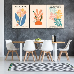 May include: Three abstract art prints featuring colorful floral and leaf designs. The prints are framed and hung on a gray wall above a wooden dining table with white chairs.