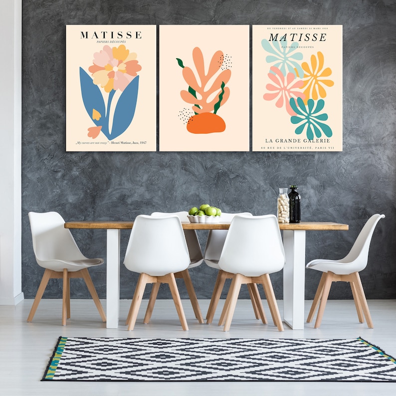 May include: Three abstract art prints featuring colorful floral and leaf designs. The prints are framed and hung on a gray wall above a wooden dining table with white chairs.