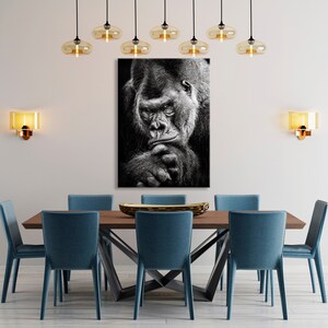 May include: Black and white photograph of a gorilla looking directly at the camera. The gorilla is sitting with its chin resting on its hand.