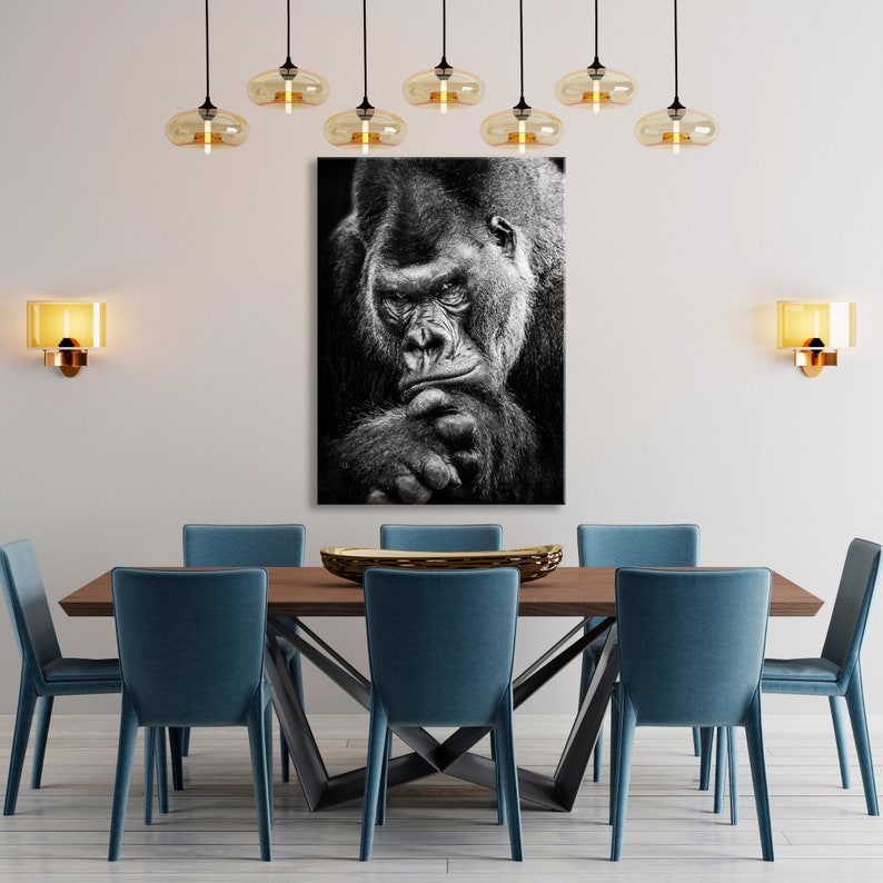 May include: Black and white photograph of a gorilla looking directly at the camera. The gorilla is sitting with its chin resting on its hand.
