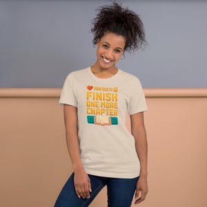 Op de afbeelding: Lichtbeige t-shirt met de tekst "Main Quest: Finish One More Chapter" in oranje en geel. Het ontwerp bevat een hart, een boek en turquoise rechthoeken. Het shirt is gemaakt van zacht materiaal.
