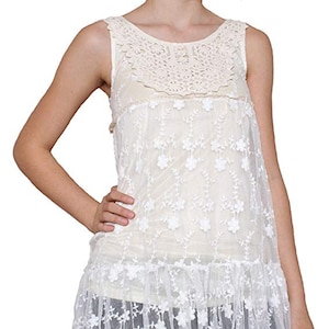 May include: Ivory-colored sleeveless tunic with a floral lace overlay. The top features a crochet neckline and a ruffled hem. The tunic is a light, airy garment, perfect for layering or wearing on its own. The design is feminine and delicate.