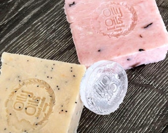 May include: Two bars of handmade soap with the Olly Oils logo. One bar is pink with black flecks and the other is beige with black seeds. A clear plastic stamp with the Olly Oils logo is also shown.