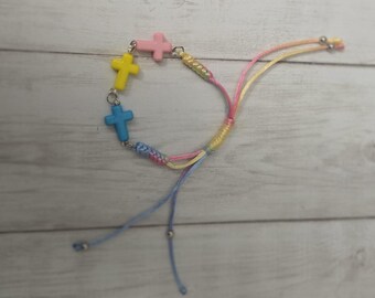 May include: A colorful bracelet featuring four cross charms in yellow, pink, and blue. The bracelet is made with a woven cord in pastel colors and adjustable sliding knots.
