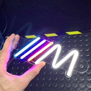 Puede incluir: Un letrero de plástico transparente con una "M" de neón blanco y tres luces LED multicolores. El letrero está siendo sostenido por una mano.