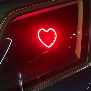 Op de afbeelding: Een rood neon hartvormig bord is zichtbaar door het achterraam van een donkerblauwe auto.