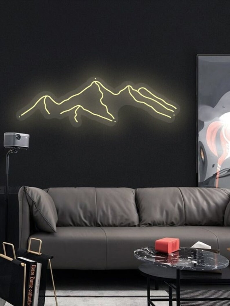 May include: A neon light sculpture of a mountain range, with a yellow glow, mounted on a black wall. The sculpture is in a living room setting with a grey sofa, a coffee table, and a projector.