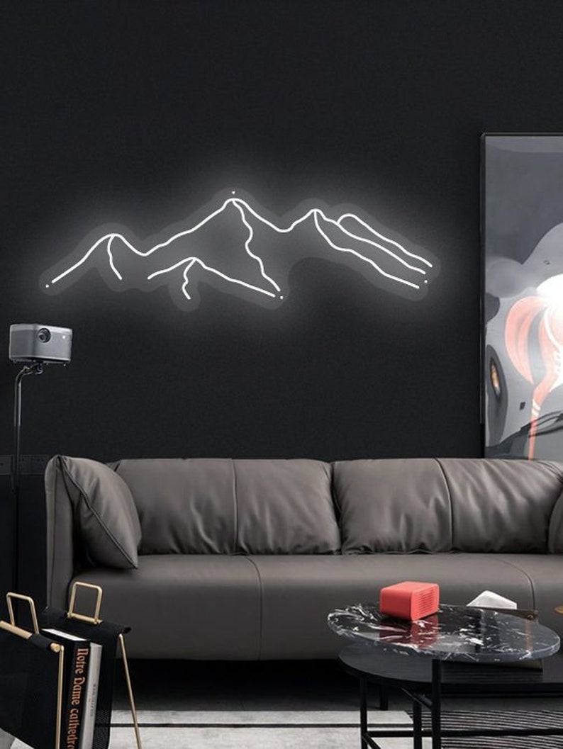 May include: A white neon sign depicting a mountain range, hanging on a black wall above a grey leather sofa. The sofa is in a living room setting with a coffee table and a black and white framed picture on the wall.