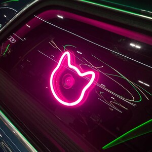 May include: A neon pink cat head outline glows against a dark background. Green and white light streaks add movement. The image has a modern, artistic feel, with a focus on the bright, stylized cat shape.