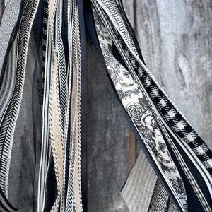 K&ouml;nnte beinhalten: Eine Sammlung dekorativer B&auml;nder in verschiedenen Mustern und Texturen, darunter Streifen, Fischgr&auml;tenmuster und florale Designs. Die B&auml;nder sind haupts&auml;chlich schwarz, wei&szlig; und beige, wodurch eine neutrale Farbpalette entsteht. Die B&auml;nder h&auml;ngen vor einem verwitterten Holzgrund.